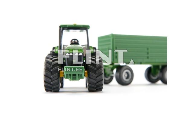 The image features a green tractor with large rubber tires and a black cabin. Behind the tractor is a green trailer with a box-like loading area.