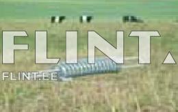 The image shows a metallic coil lying on a grassy pasture. In the background, there are green fields with some black and white cows.